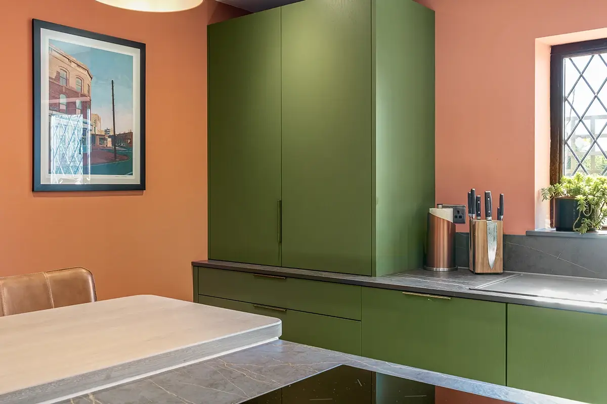 Green kitchen units and worktop against terracotta walls with framed artwork