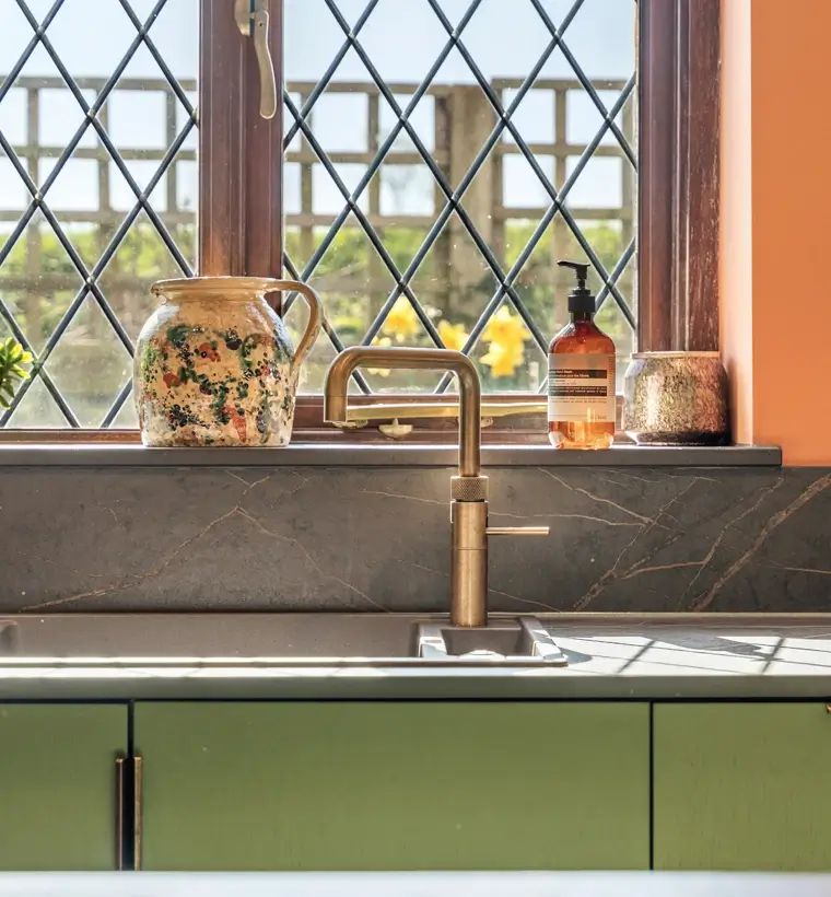 Kitchen sink with brass-coloured tap beneath leaded windows and green cabinets